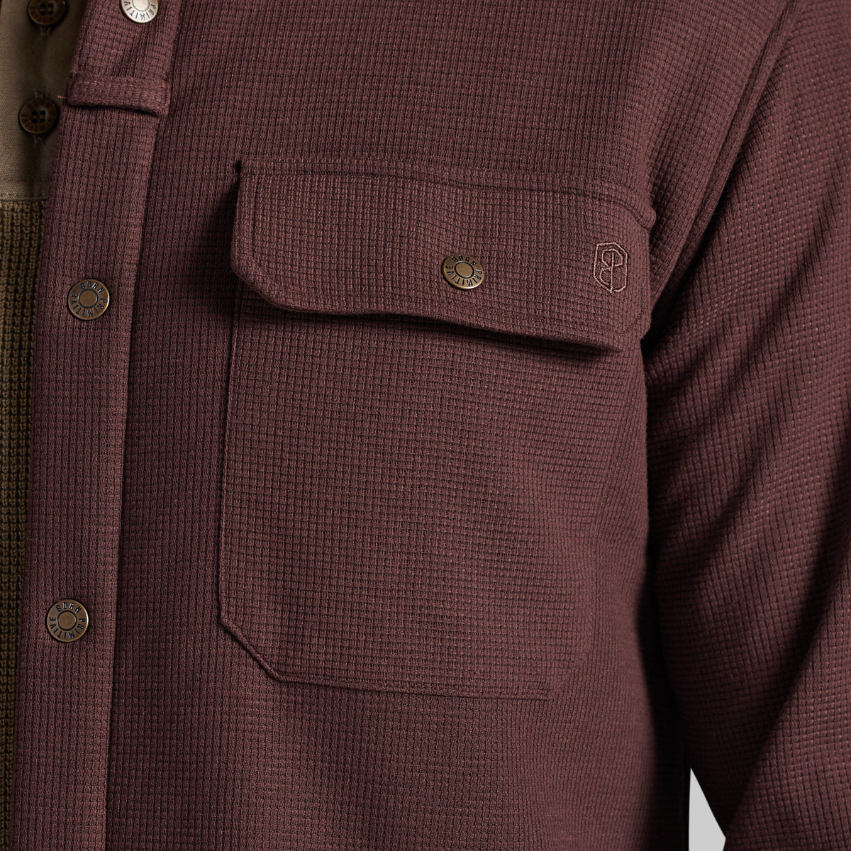 Close-up of a waffle-knit button-front shirt jacket in a muted burgundy-brown color, featuring a chest pocket with a flap and metal snap, plus a small embroidered logo.