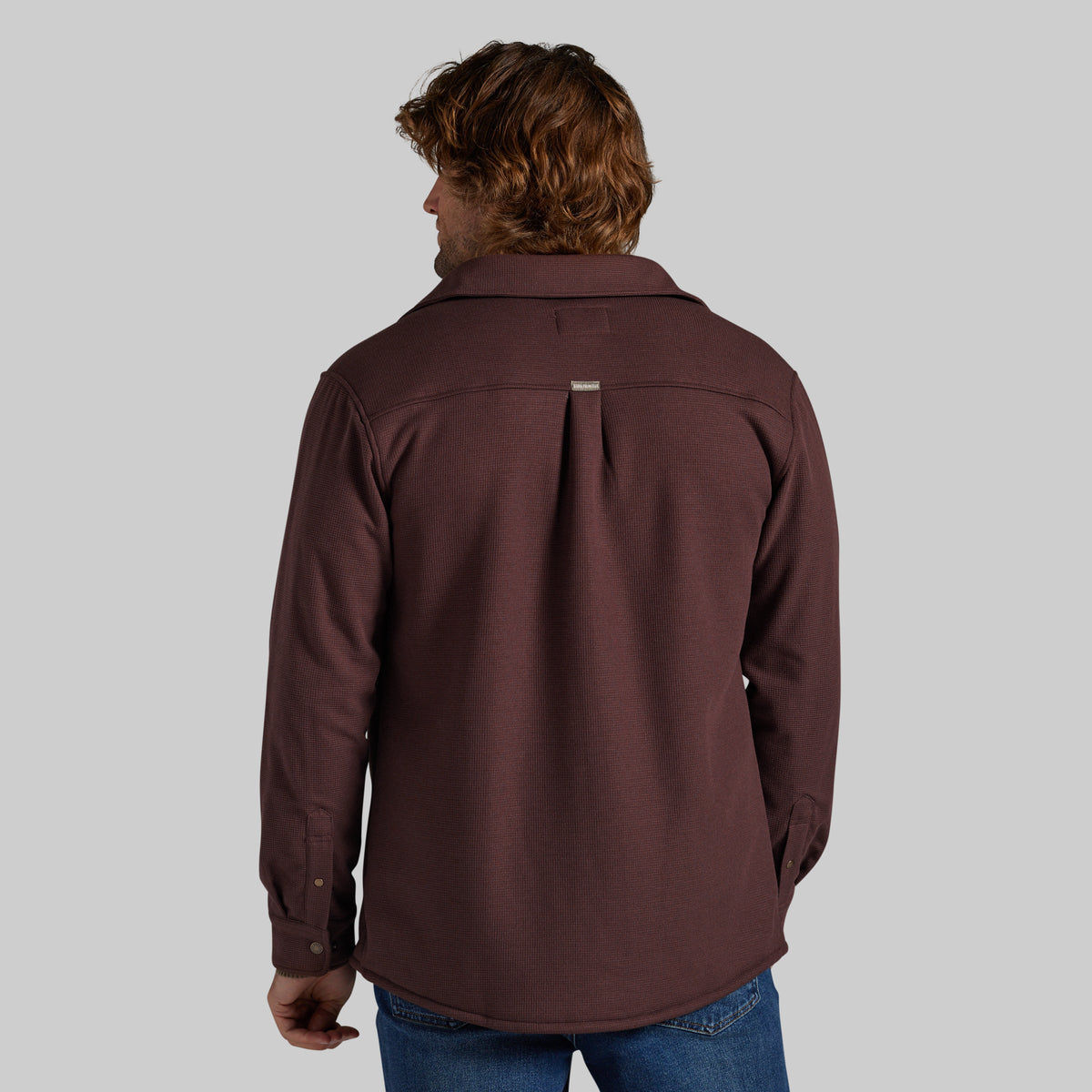 A man viewed from the back wearing a dark brown long-sleeve button-up jacket and blue jeans, set against a solid gray studio background.