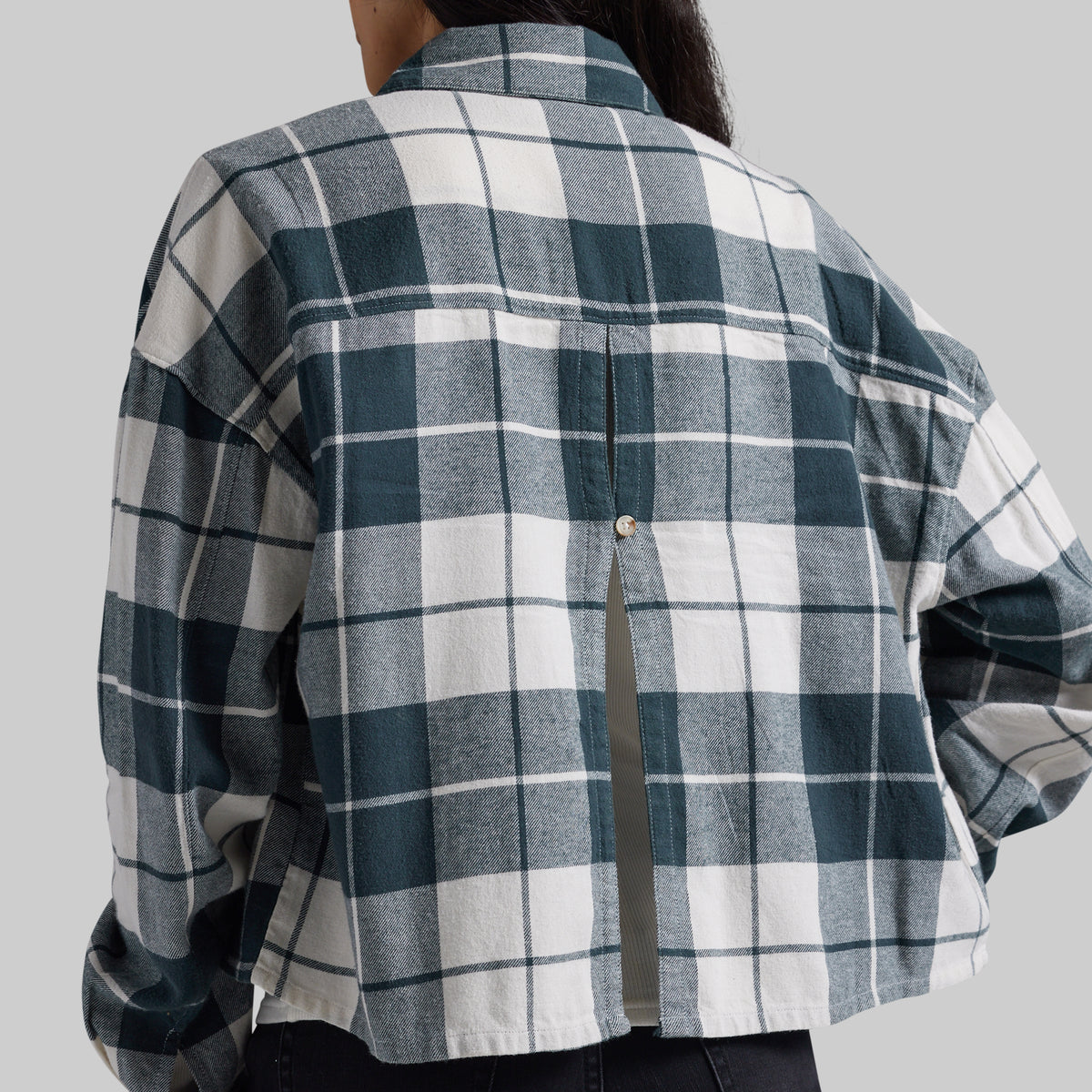 A person shown from the back wearing a plaid button-up shirt in dark teal, white, and gray, with a small button closure at the back. The background is a solid light gray studio backdrop.