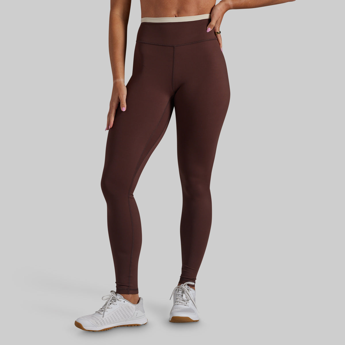 A model wearing dark brown high-waisted leggings and white athletic sneakers, shown from the waist down against a solid light gray background.