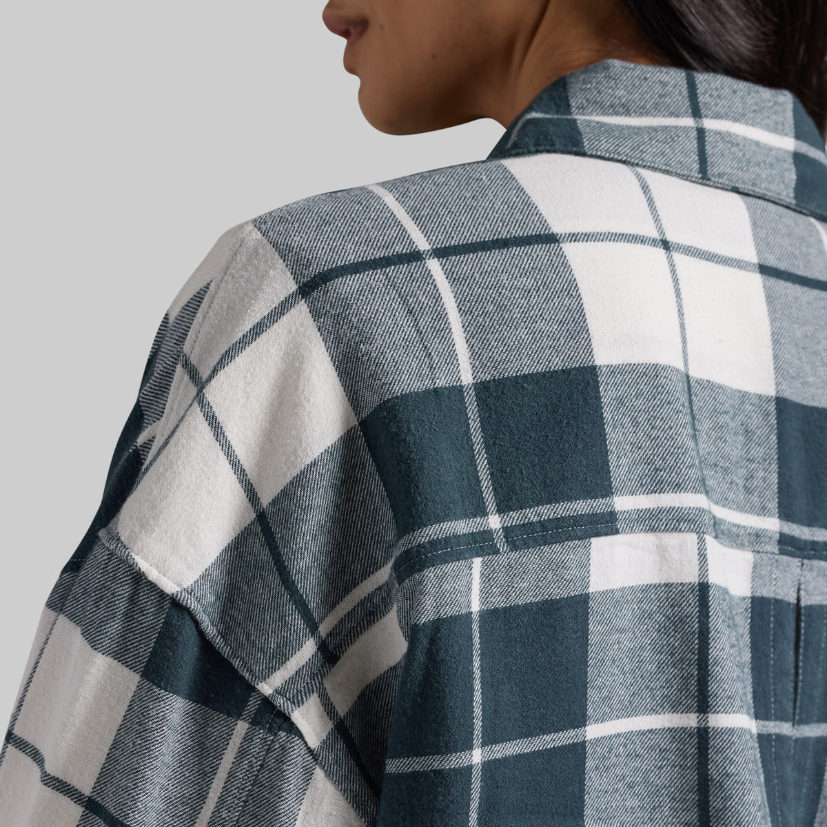 Close-up of a person wearing a plaid flannel shirt, showing the back and shoulder area with a dark teal, gray, and white check pattern against a light gray background.