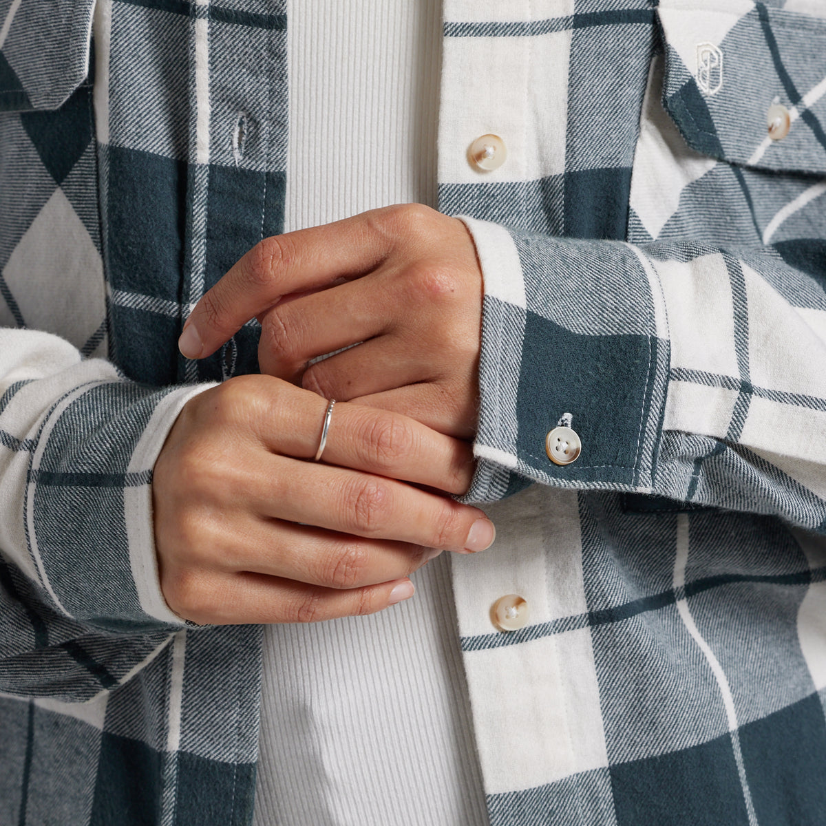 Close-up of a person wearing a blue and white plaid button-up shirt over a white ribbed knit top, with both hands visible adjusting the cuff.