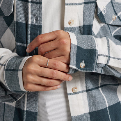 Close-up of a person wearing a blue and white plaid button-up shirt over a white ribbed knit top, with both hands visible adjusting the cuff.