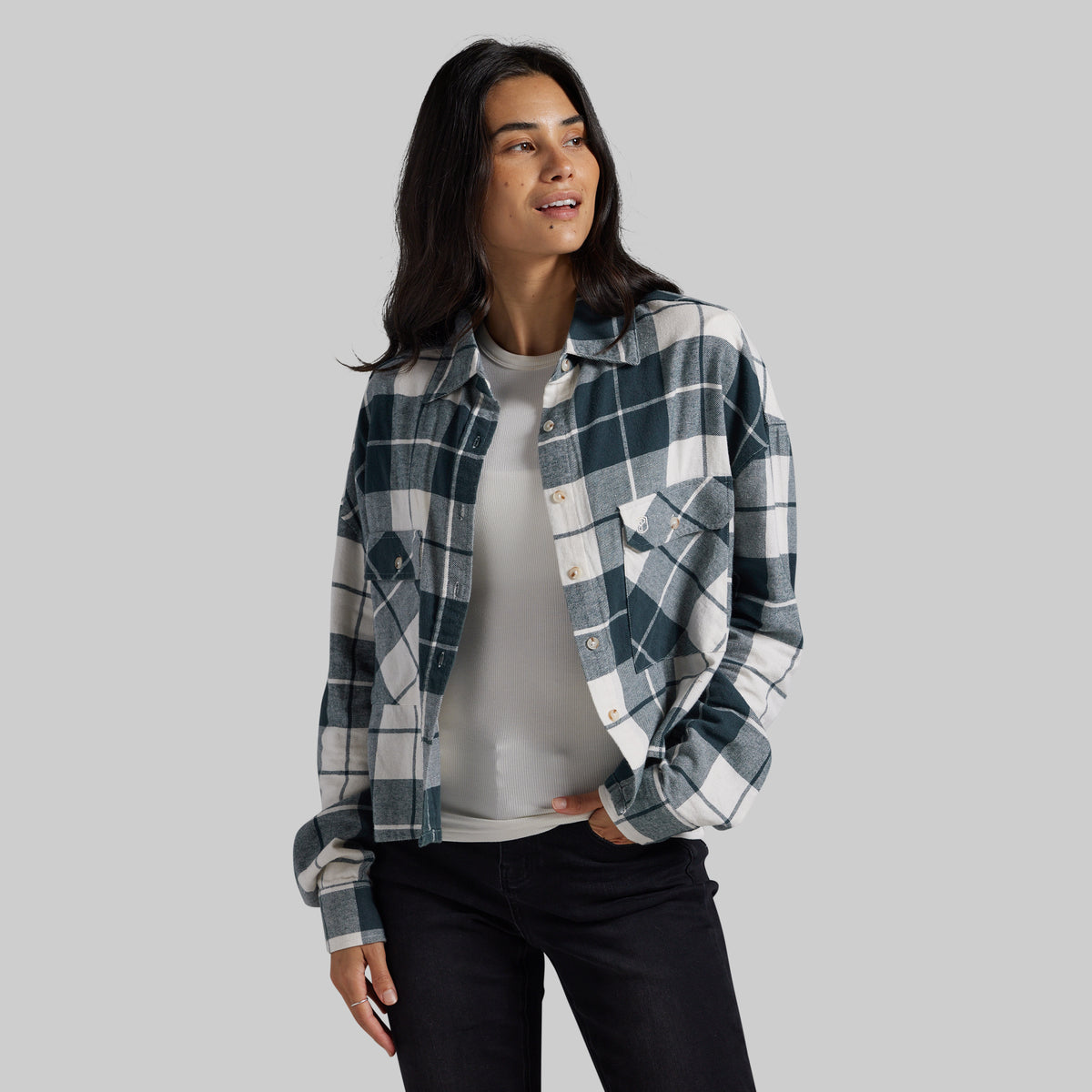 A woman modeling a dark teal and white plaid flannel shirt over a light gray t-shirt and black jeans, set against a solid gray studio background.