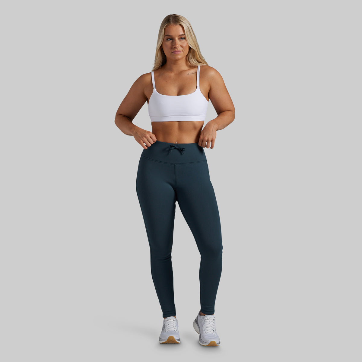 A woman modeling high-waisted athletic leggings in a dark teal color, paired with a white sports bra and white sneakers, against a solid light gray background.