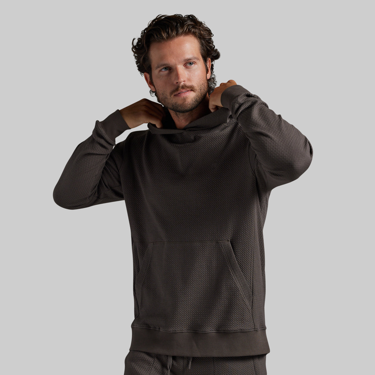 A man wearing a dark grey textured pullover hoodie with a high neck and kangaroo pocket, posing with his hands behind his neck against a solid gray background.
