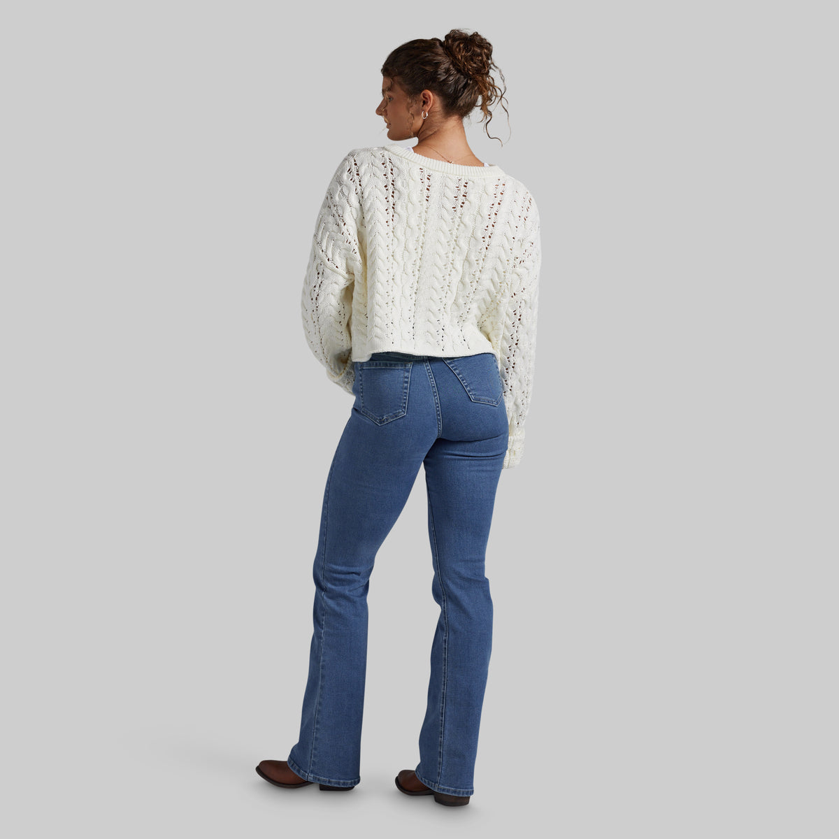 A woman shown from the back wearing a cream cable-knit cropped sweater and blue jeans, standing against a solid gray studio background.