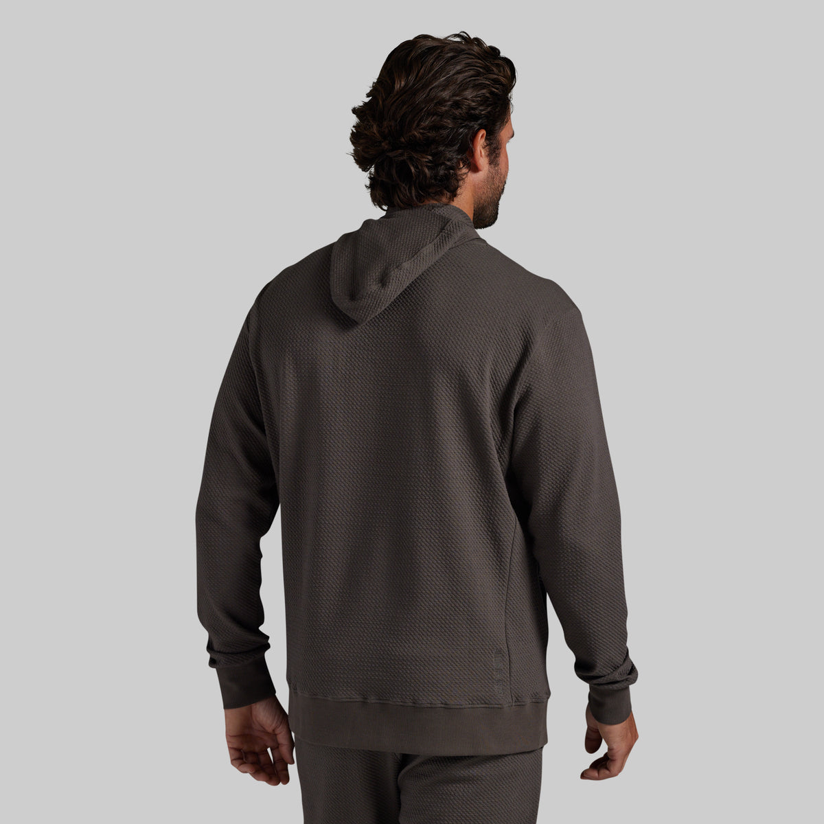 A man viewed from behind wearing a dark grey textured hoodie and matching pants against a solid gray background.