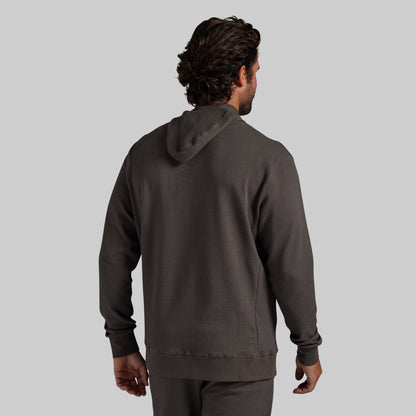 A man viewed from behind wearing a dark grey textured hoodie and matching pants against a solid gray background.