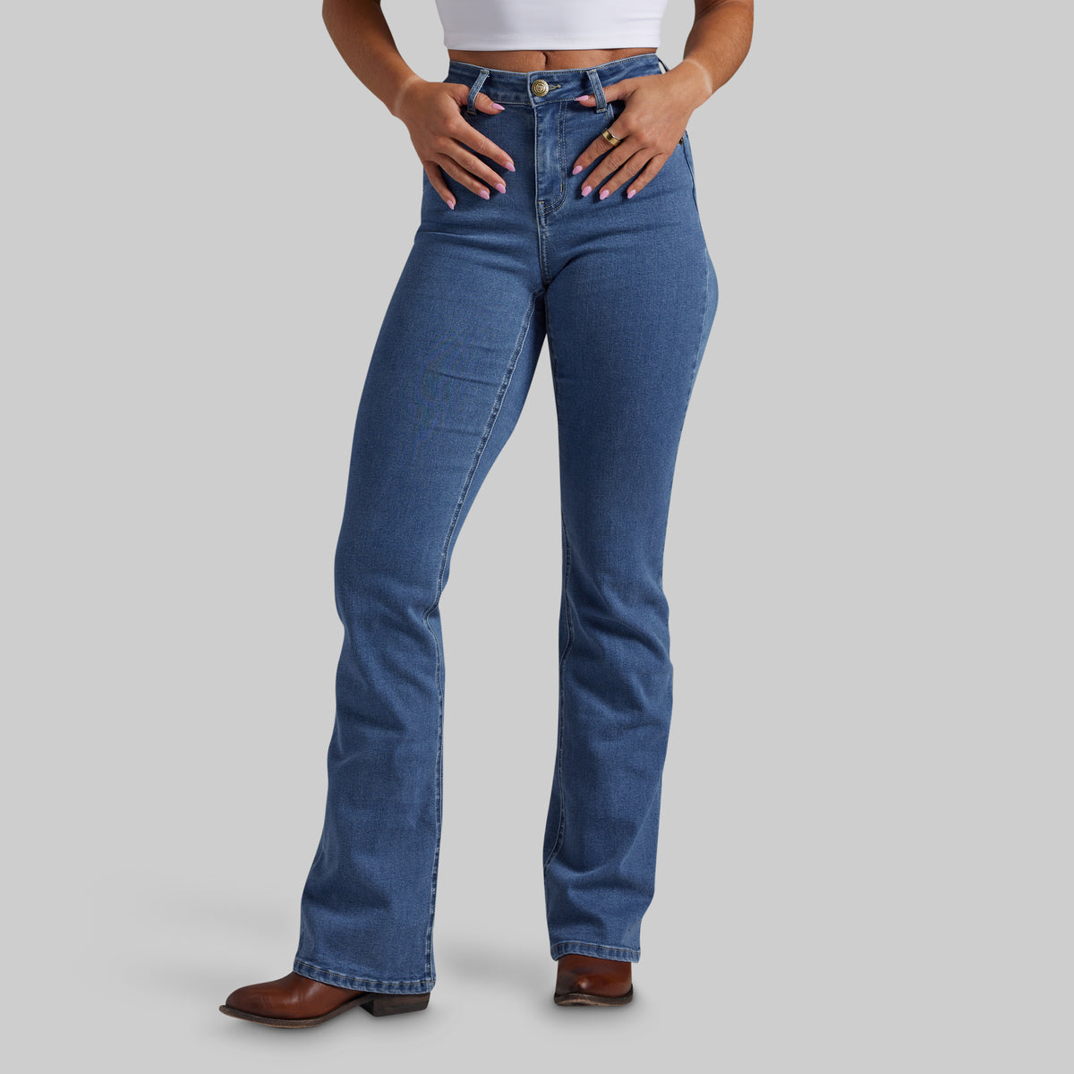 A model wearing a pair of high-waisted, bootcut blue denim jeans with a faded wash, paired with a white crop top and brown leather boots, set against a solid gray studio background.