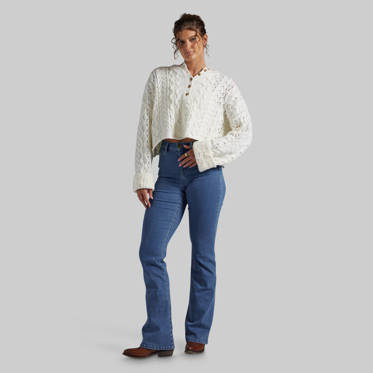 A woman modeling a white cable-knit cropped button-up shirt with blue flared jeans and brown boots, standing against a solid gray studio background.