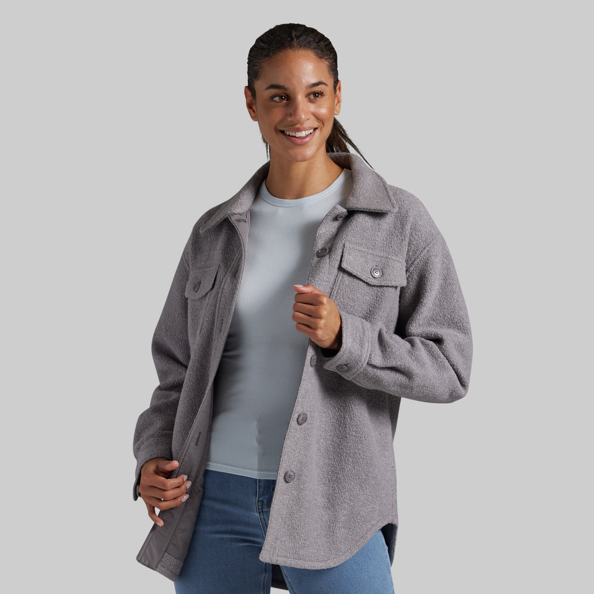 A woman wearing a heather gray button-up jacket over a light blue top and blue jeans, standing against a solid gray studio background.
