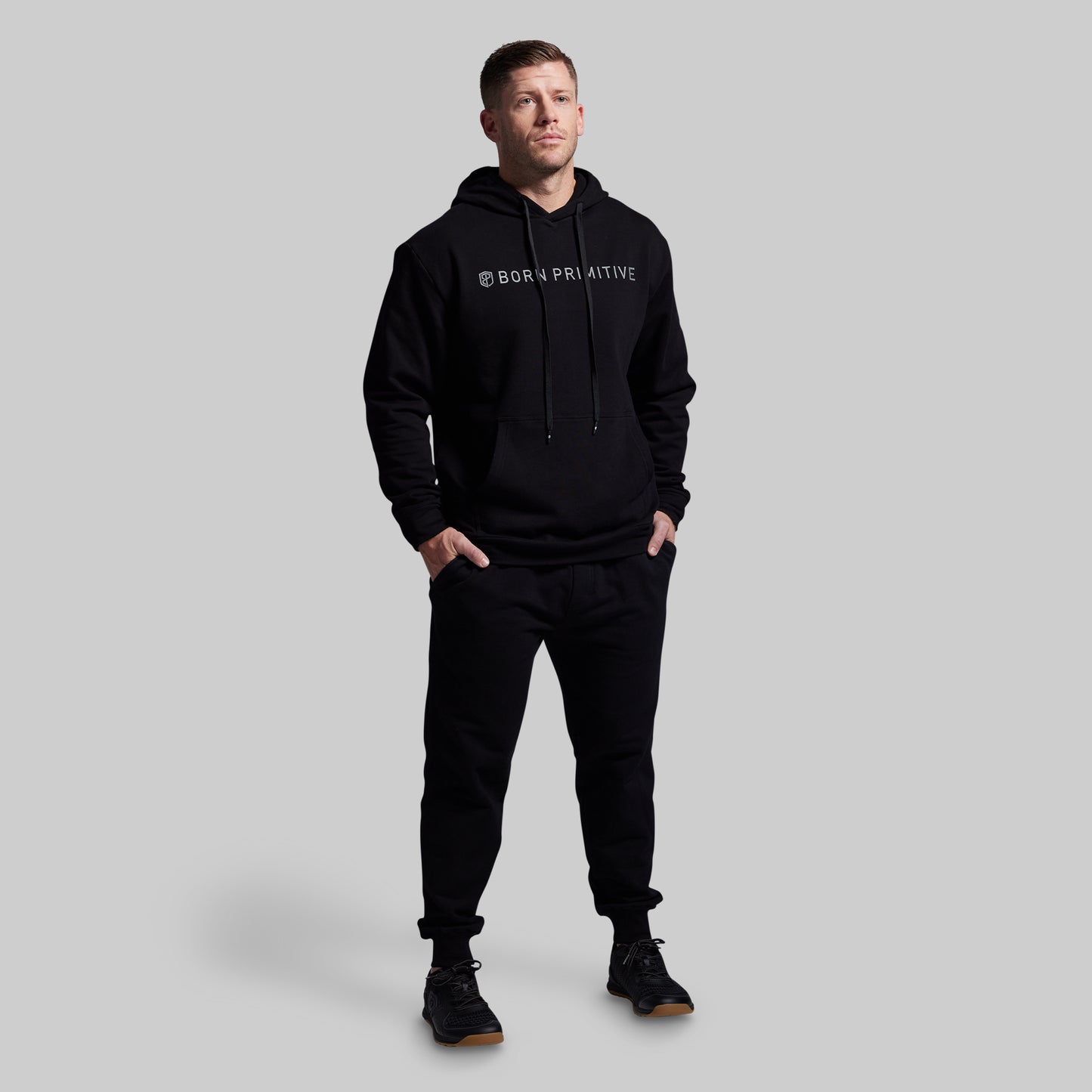 Man wearing a black hoodie and pants with a brand logo on a gray background