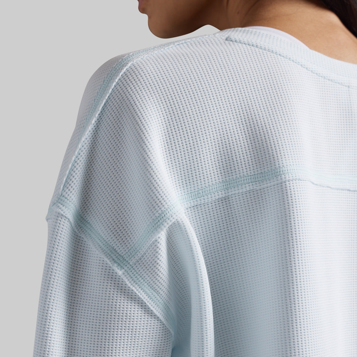 Close-up of a person wearing a light icy blue waffle-knit long-sleeve top, showing the textured fabric and shoulder seam details against a neutral gray background.