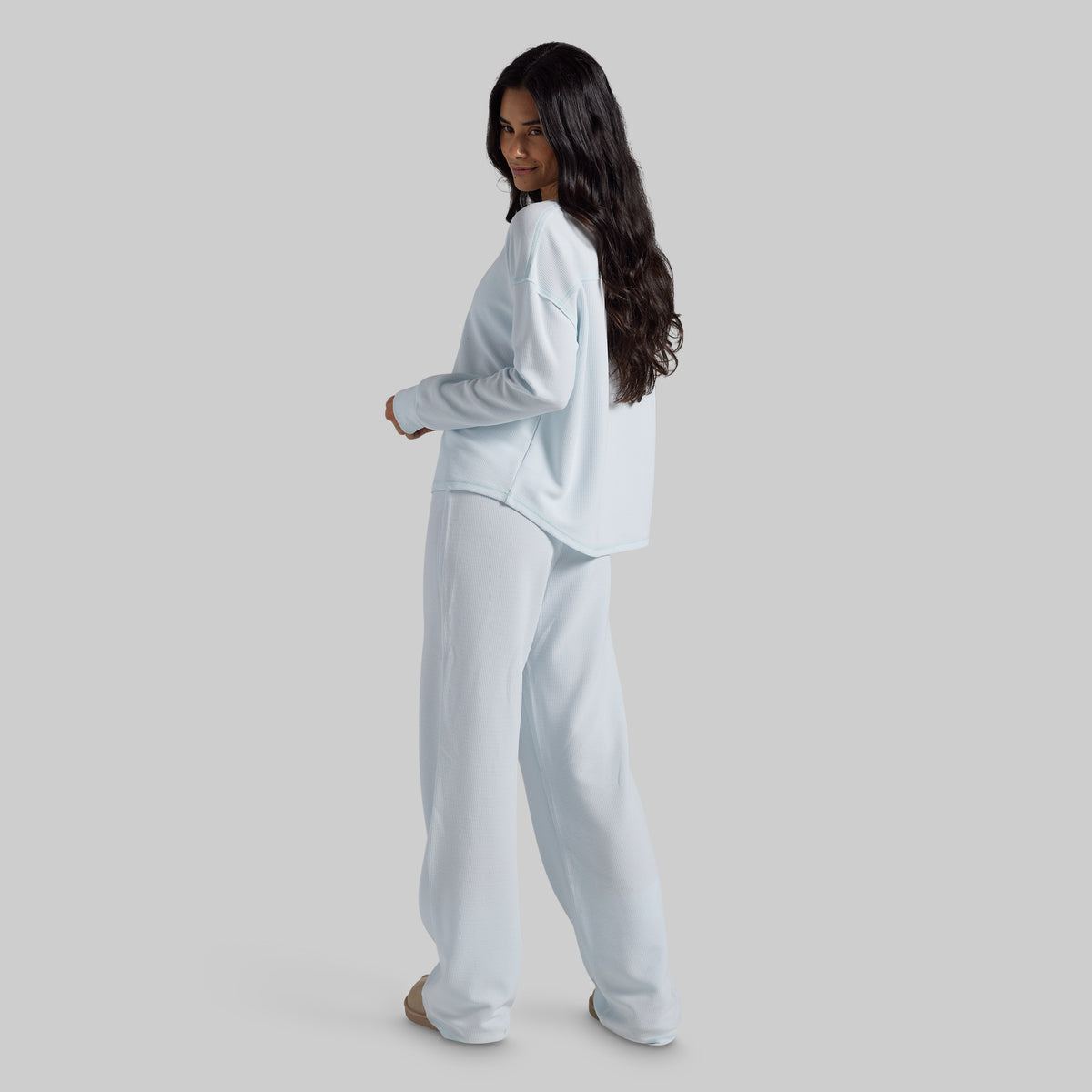 A woman modeling a light blue lounge set consisting of a long-sleeve top and matching wide-leg pants, shown from a side-back angle against a solid gray studio background.