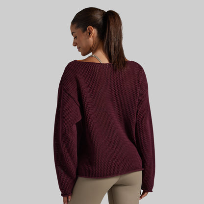 A woman shown from the back wearing a textured, wide-neck knit sweater in deep burgundy, paired with beige leggings, against a solid light gray background.