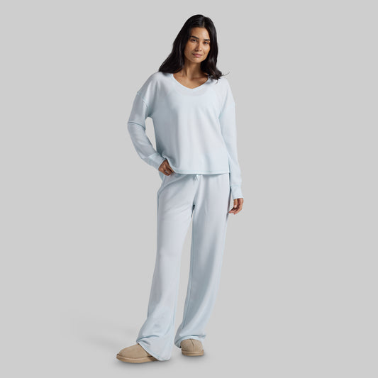 A woman modeling a light blue two-piece loungewear set consisting of a long-sleeve V-neck top and matching wide-leg pants, paired with beige slippers against a solid gray studio background.