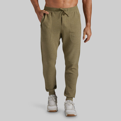 A pair of men's olive green textured jogger pants with a drawstring waist, side pockets, and ribbed ankle cuffs, shown on a model from the waist down against a light gray background.