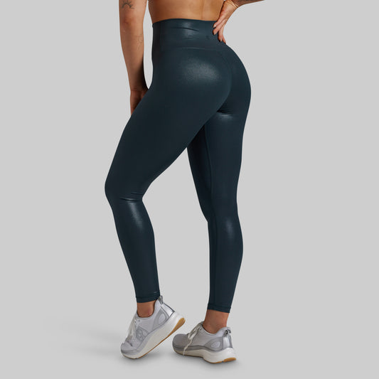 A person wearing high-waisted, shiny dark teal leggings, shown from the back and side against a light gray studio background. The model is also wearing light gray sneakers.