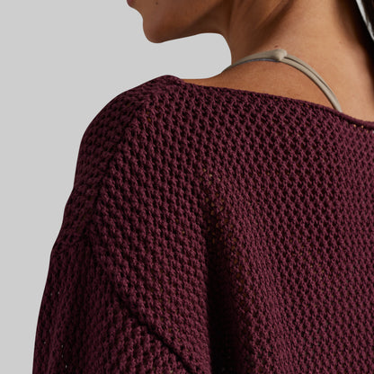 Close-up of a person wearing a chunky knit burgundy sweater, showing the textured fabric and shoulder strap detail against a light gray background.