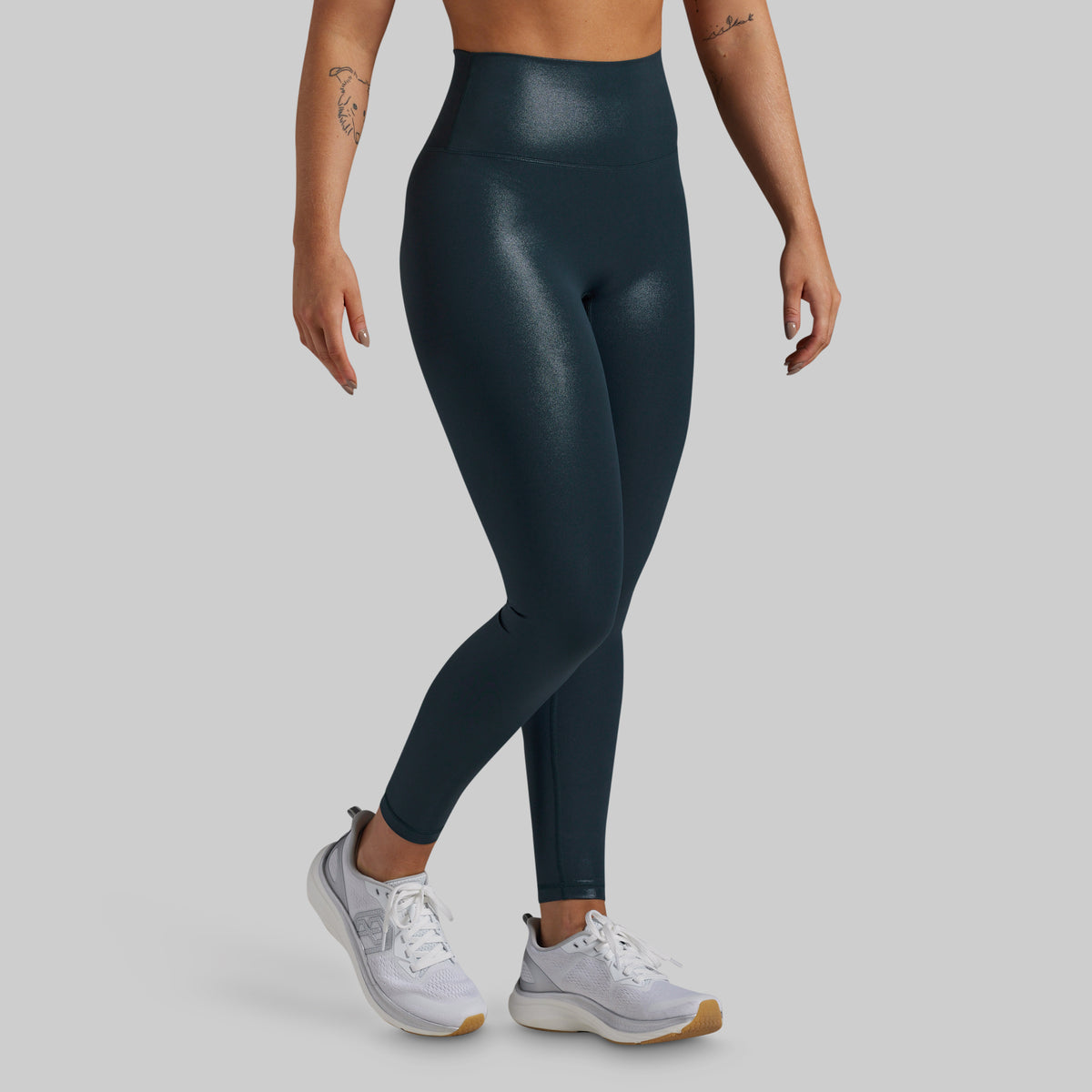 A model wearing high-waisted, shiny dark teal athletic leggings and white sneakers, shown from the waist down against a solid gray background.