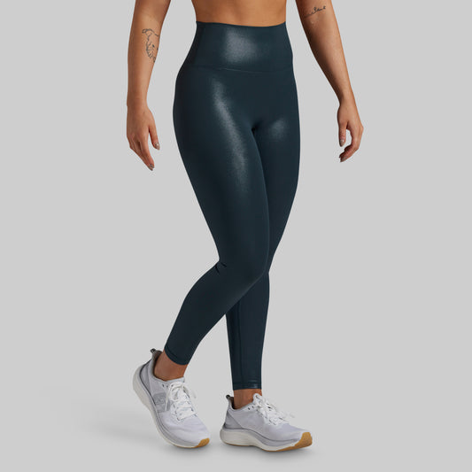 A model wearing high-waisted, shiny dark teal athletic leggings and white sneakers, shown from the waist down against a solid gray background.