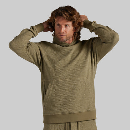 A man wearing a textured olive green hooded sweatshirt and matching sweatpants, posing with his hands behind his head against a solid gray background.