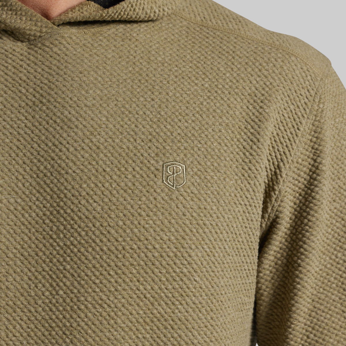 Close-up of a textured olive green hoodie with a small white embroidered shield logo on the chest, worn by a model.