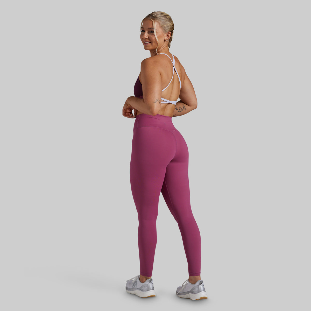 A woman modeling high-waisted, form-fitting leggings in a dusty rose pink color, paired with a white sports bra and gray sneakers, shown from a three-quarters back view against a solid gray studio background.