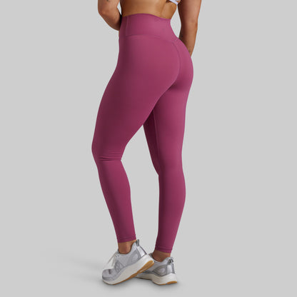 A model from the back wearing high-waisted, form-fitting leggings in a dusty magenta pink color, paired with white and silver sneakers, against a solid light gray background.