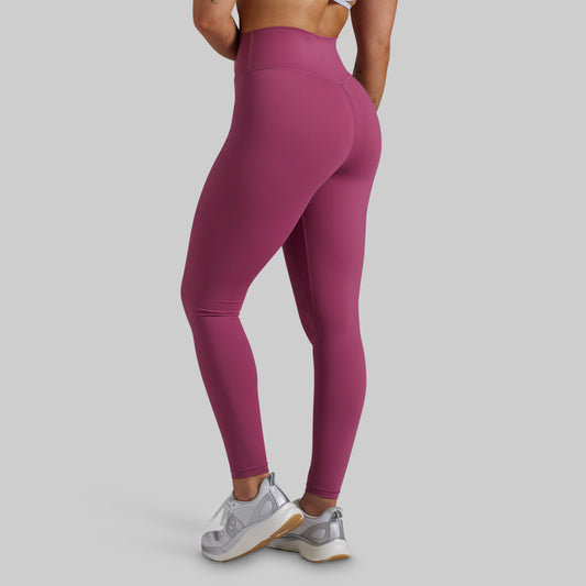 A model from the back wearing high-waisted, form-fitting leggings in a dusty magenta pink color, paired with white and silver sneakers, against a solid light gray background.