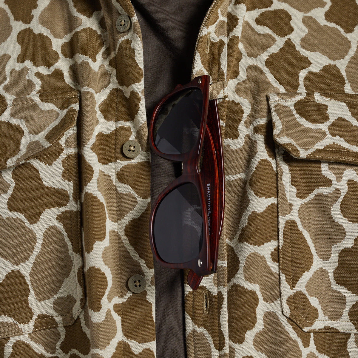 A close-up of a brown and beige frog camo flannel with a dark button-down shirt underneath and a pair of sunglasses hanging from the placket.