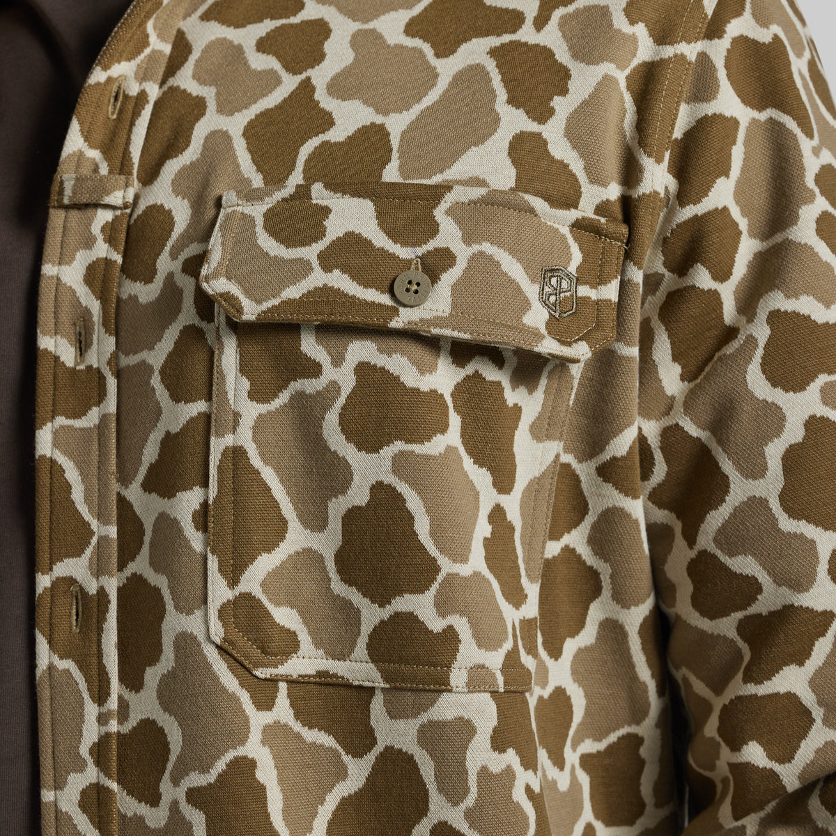 Close-up of a brown and beige frog camo featuring a buttoned chest pocket and a small embroidered logo.