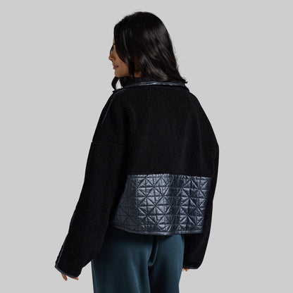 A woman shown from the back wearing a black cropped jacket with a dark gray quilted geometric patterned panel on the lower back, paired with dark teal pants, against a solid light gray background.