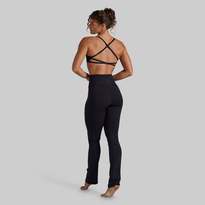 A woman is shown from the back wearing a black strappy athletic bra and black high-waisted yoga pants against a solid gray background.