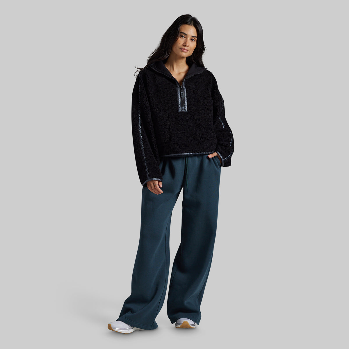 A woman modeling a black fleece half-zip pullover with textured knit sleeves and dark teal wide-leg sweatpants, standing against a solid gray studio background.