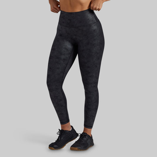 A model wearing high-waisted, full-length athletic leggings with a dark charcoal gray and black mottled pattern, paired with black athletic shoes, against a solid light gray background.