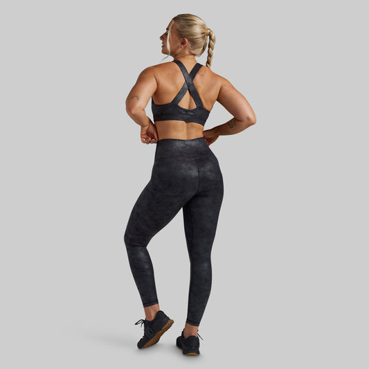 A woman shown from the back wearing a matching dark gray and black athletic set, featuring a sports bra with crisscross straps and high-waisted leggings with a subtle mottled pattern, set against a solid gray background.