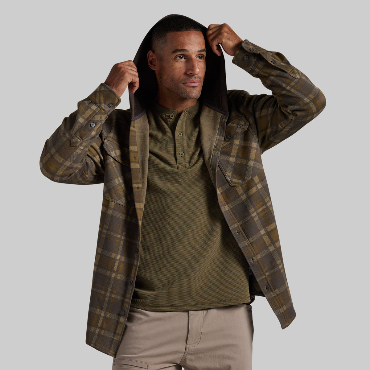 A man wearing a brown and olive green plaid hooded flannel shirt over an olive green henley shirt and khaki pants, posing with the hood up against a solid gray background.