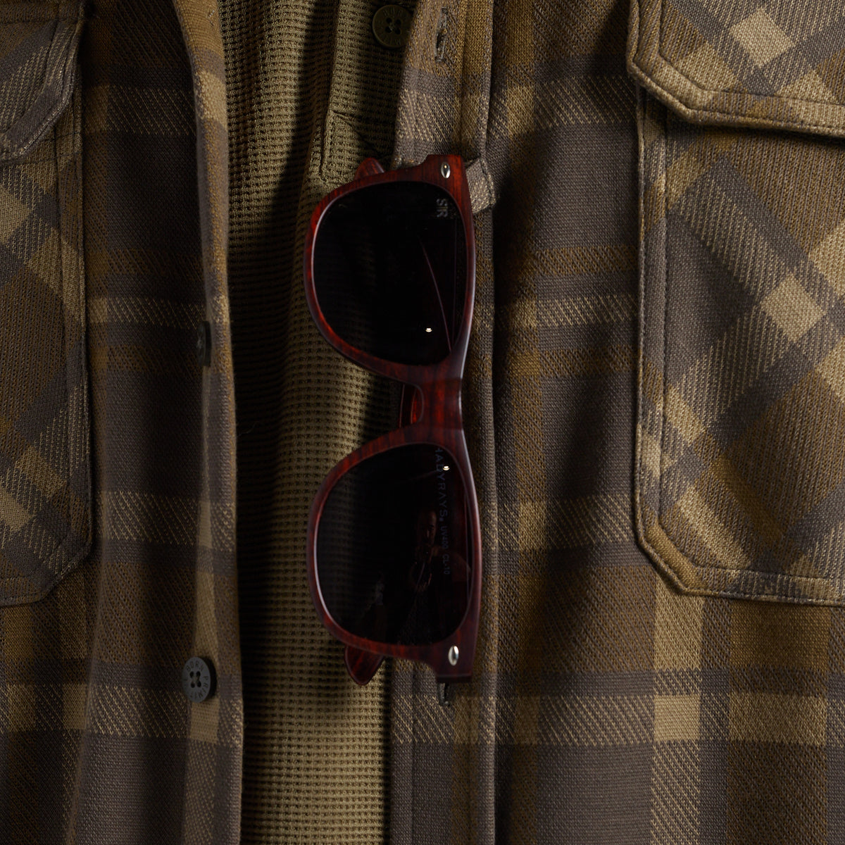 Close-up of sunglasses hanging from the sunglass loop of a brown and tan plaid flannel shirt.