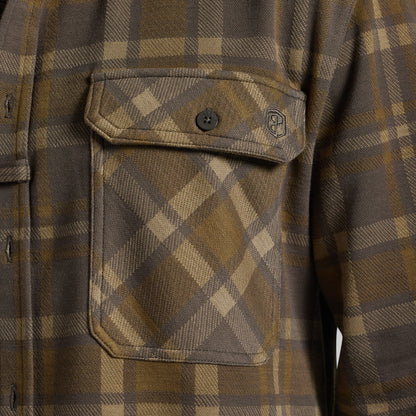 Close-up of a brown and tan plaid flannel shirt pocket with a button closure and a small geometric logo on the chest flap.