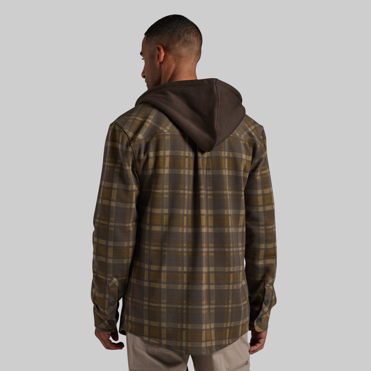 A man viewed from behind wearing a brown and olive green plaid hooded overshirt with a dark brown hood, paired with light khaki pants, against a solid gray background.