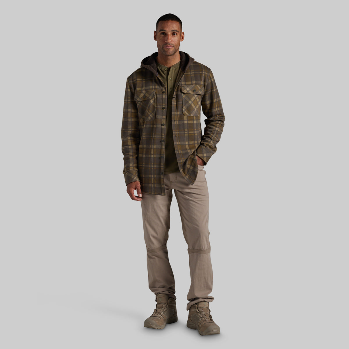 A man modeling a brown and olive green plaid hooded flannel shirt jacket, styled with a dark olive t-shirt, beige cargo pants, and brown boots, against a solid gray background.