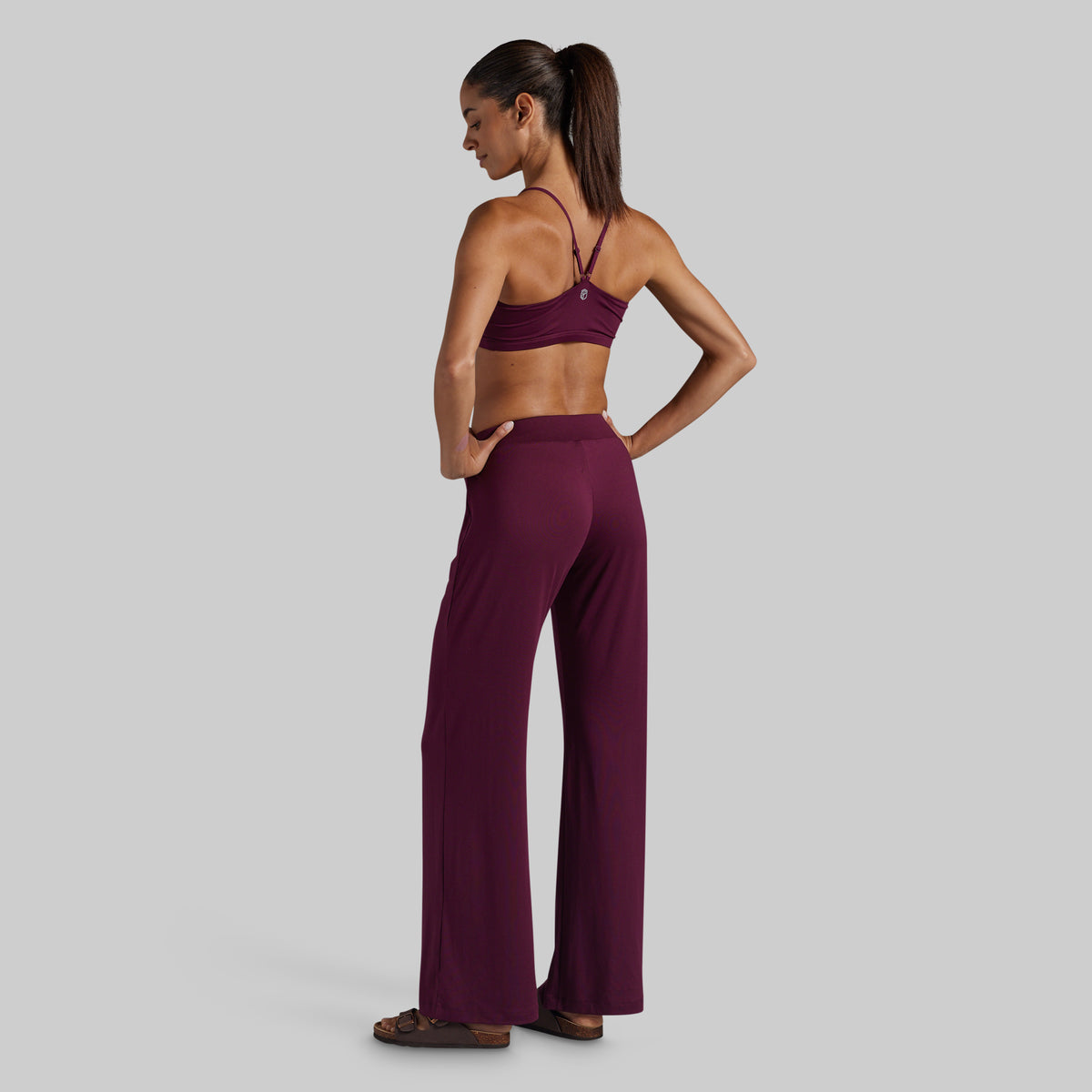 A woman stands in profile wearing a matching burgundy activewear set consisting of a sports bra and high-waisted wide-leg pants, photographed against a solid light gray background.