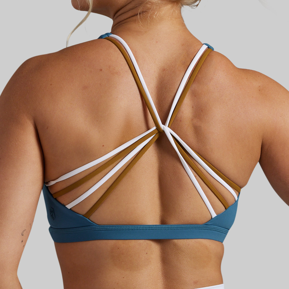 Rear view of a woman wearing a teal sports bra with multiple thin crossover straps in white and mustard brown on a light gray background.