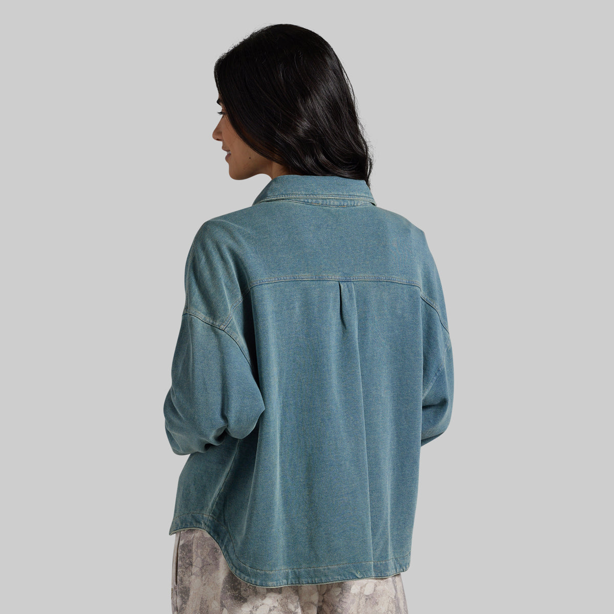 A woman shown from the back wearing an oversized blue denim shirt with a button-down collar and long sleeves, photographed against a solid light gray background.