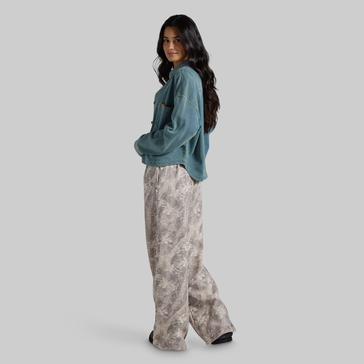 A woman modeling a teal cropped denim jacket and wide-leg beige and gray patterned pants against a solid light gray background.