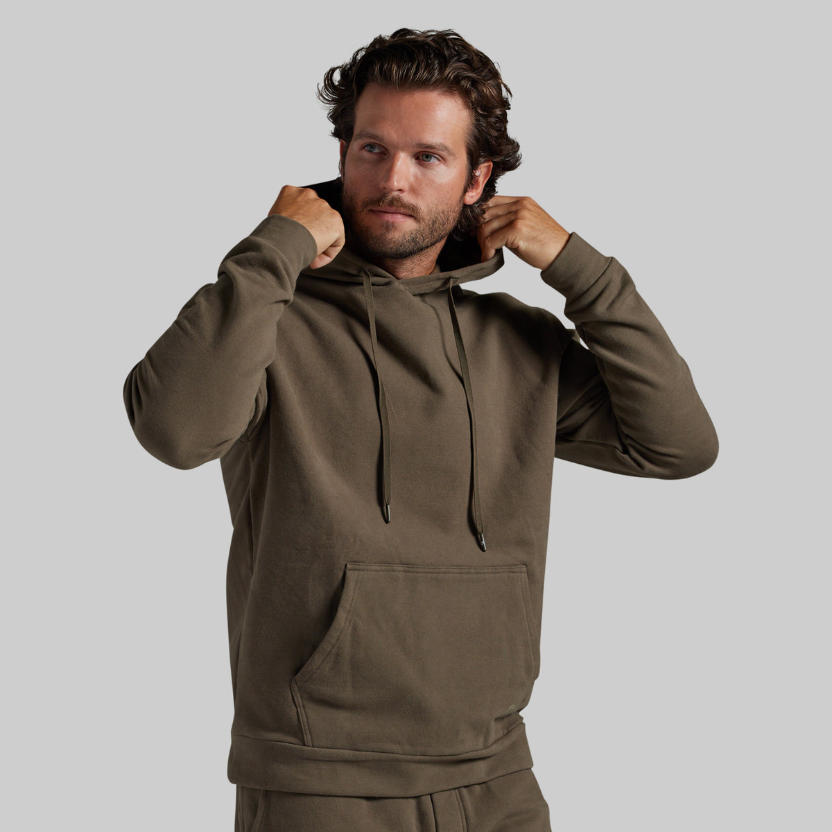 A man wearing an olive brown pullover hoodie with matching sweatpants, posing with his hands on the hood drawstrings against a solid gray background.