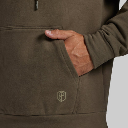 Close-up of a person wearing an olive brown pullover hoodie, showing a hand in the front pocket with a small embroidered shield logo near the lower pocket.