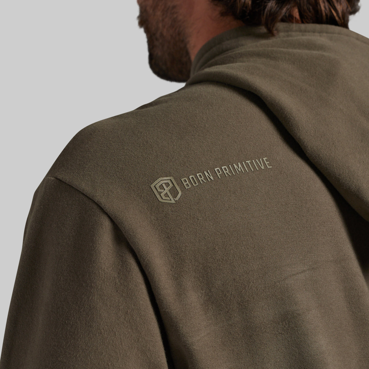 Close-up of a person wearing an olive brown hooded sweatshirt, showing the back of the neck, shoulder, and upper back. The brand name 'BORN PRIMITIVE' with its geometric logo is printed in tonal olive on the upper left shoulder.